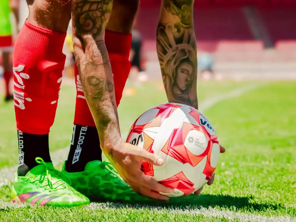 Anonymous close-up of a football on wet grass near the penalty spot, stadium seats blurred in background