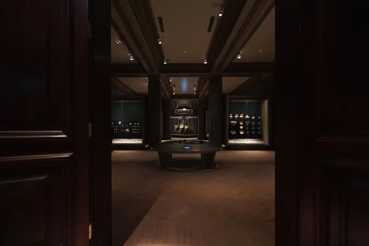 A dimly lit gallery interior with rows of black-and-white photographs on white walls, an empty bench in the middle, soft directional light highlighting frames