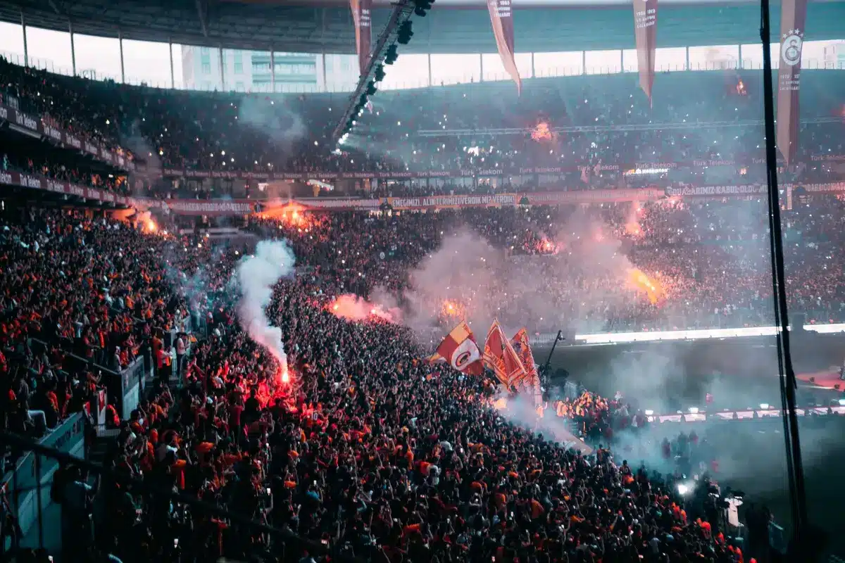 Empty away section at a small stadium with scattered anonymous supporters, scarves and flags blurred, autumn evening build-up