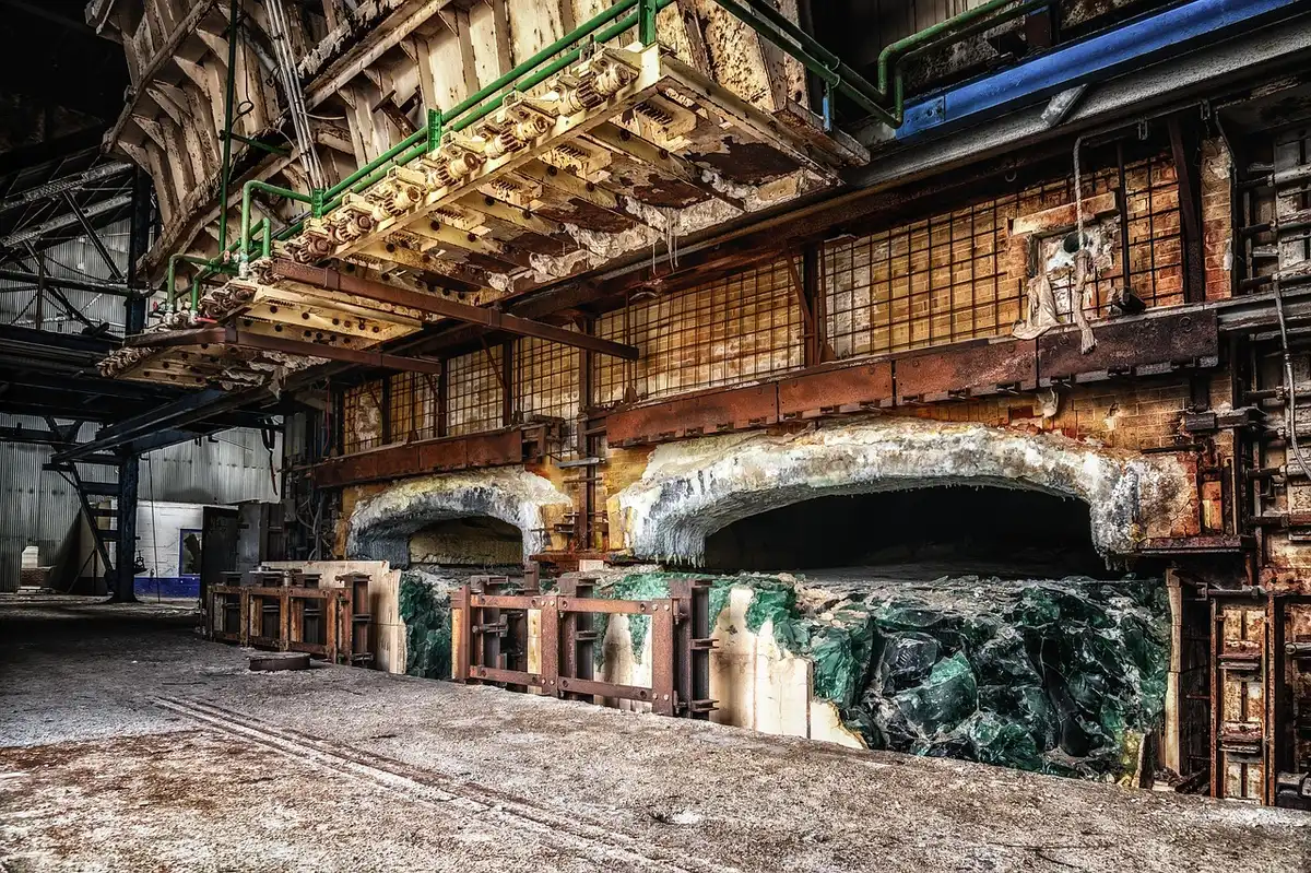 Wide shot of an overgrown factory courtyard with cracked concrete, scattered pallets and faint path toward a distant warehouse