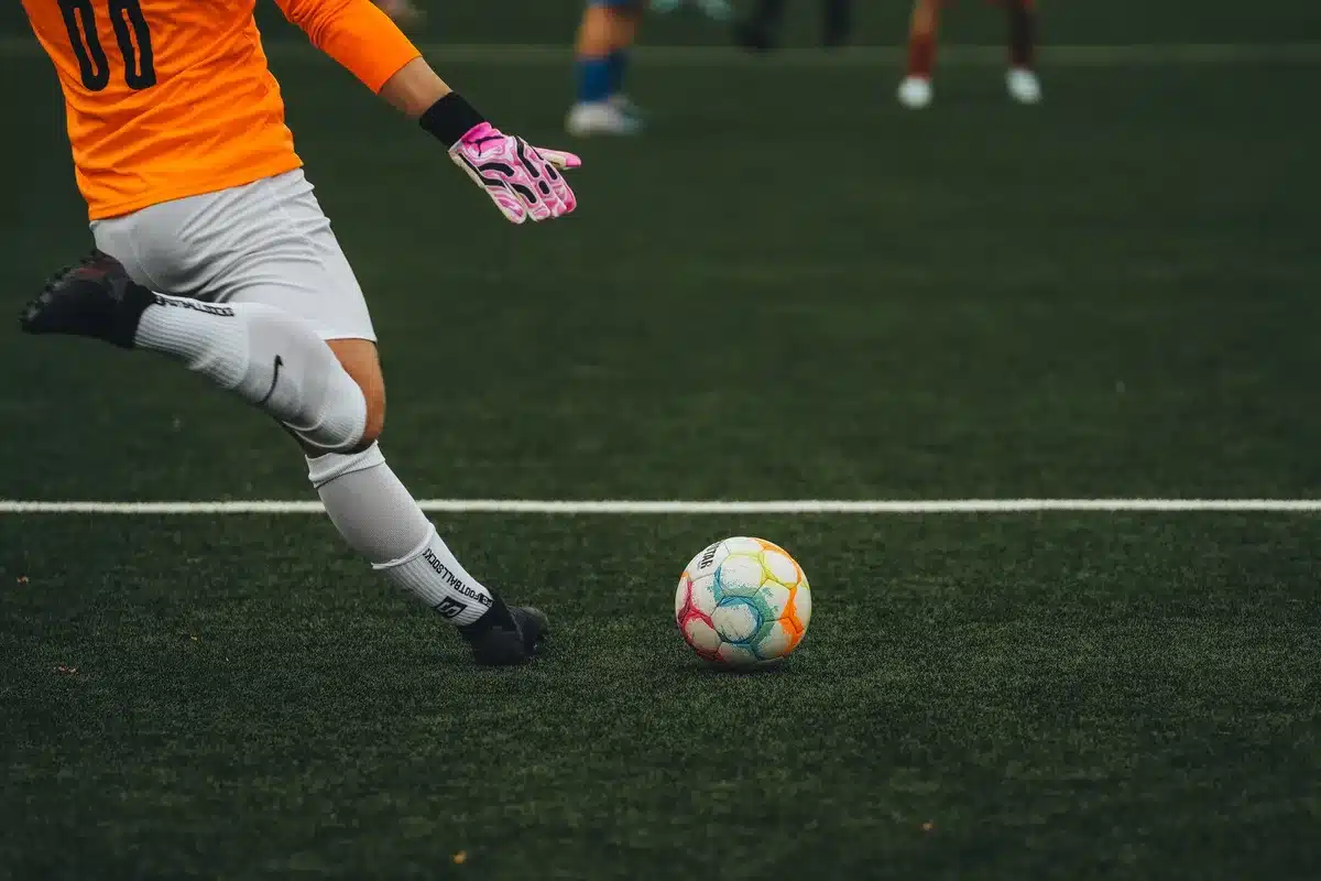 An empty football pitch under stadium lights with a lone match ball on the penalty spot, wet grass reflecting light