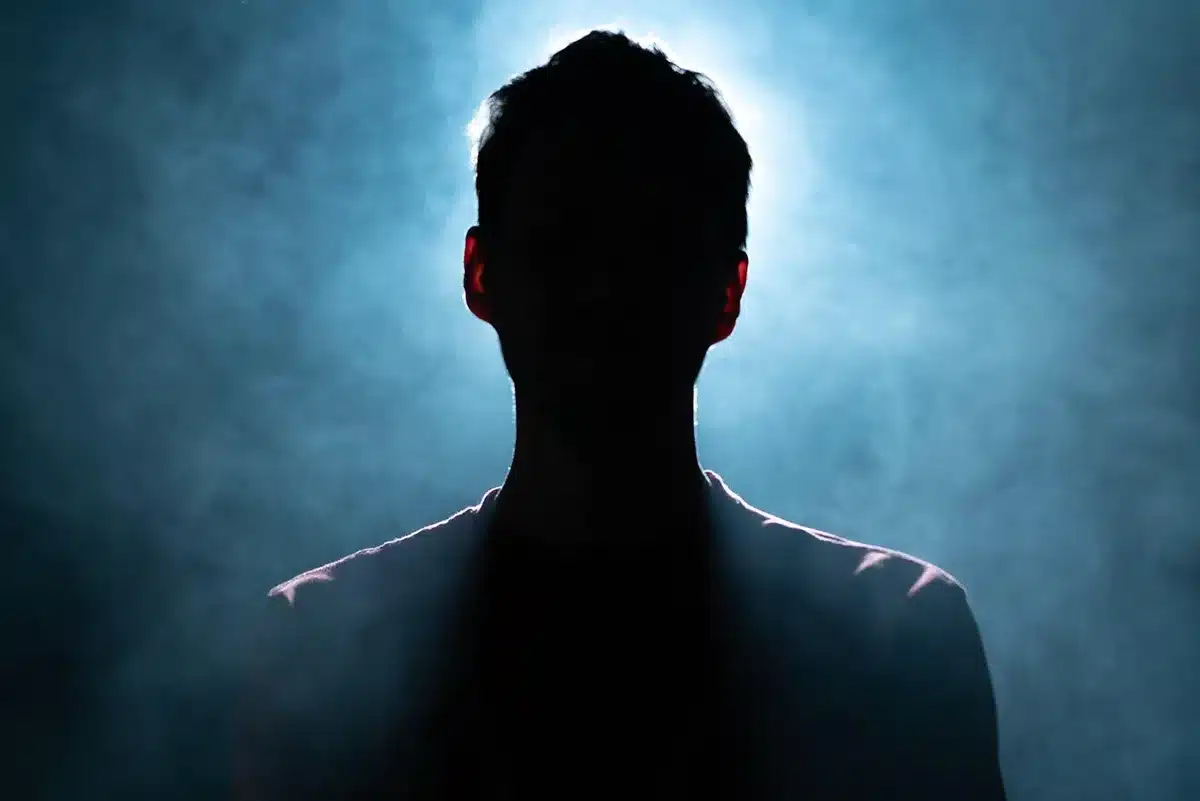 Wide shot of an anonymous young singer on a dimly lit stage, backlit silhouette holding a microphone stand, empty blurred audience seats beyond — evokes audition tension