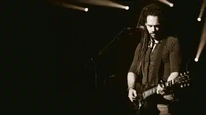 Mani che tengono un vinile davanti a una chitarra su un palco vuoto, luci di scena sfocate